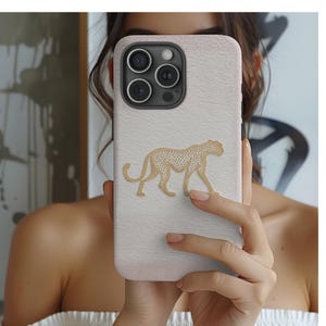 May include: A light pink and white phone case with a gold cheetah design. The case has a textured surface and a camera with three lenses. The cheetah is walking with its head up.