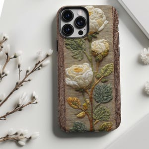 Könnte beinhalten: Eine Handyhülle mit einem floralen Design mit weißen Rosen, grünen Blättern und einem braunen Rand. Die Hülle hat ein strukturiertes Aussehen, wobei die Blumen und Blätter erhaben erscheinen.