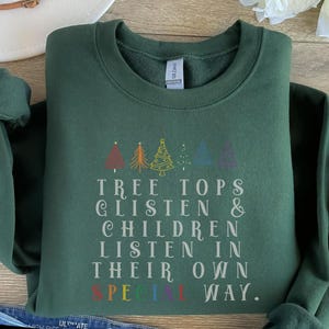 Op de afbeelding: Een donkergroen sweatshirt met een kerstthema. De tekst luidt "Tree Tops Glisten & Children Listen In Their Own Special Way". Het ontwerp bevat een rij gestileerde kerstboom met regenboogkleurige accenten.
