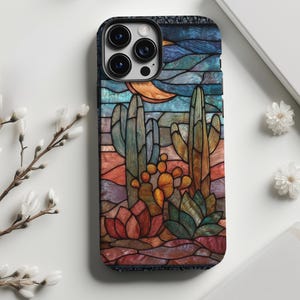 May include: A phone case featuring a desert landscape design. The case depicts cacti, a crescent moon, and a colorful sky in a stained-glass style. The case has a black border and a glossy finish, offering protection and style.