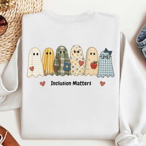 May include: A white crewneck sweatshirt featuring a graphic of six ghosts with different patterns, including notebook paper, yellow stripes, green plaid, floral, ruler markings, and blue gingham, with the text Inclusion Matters and two hearts.