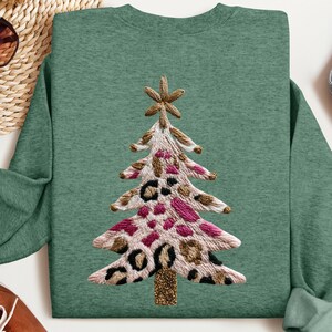 May include: A green sweatshirt featuring an embroidered Christmas tree design. The tree has a star on top and is decorated with pink, black, and brown leopard print spots. The tree trunk is gold glitter.