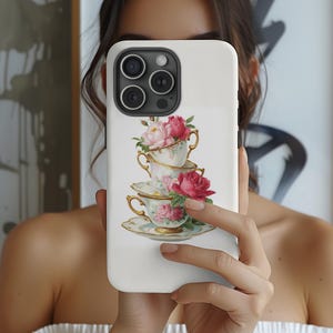 May include: White phone case featuring a stack of three teacups and saucers with a floral design. The teacups are adorned with pink roses and peonies, and have gold trim. The case is held in front of a person.