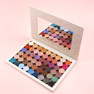 May include: An open white eyeshadow palette with a geometric design and a mirror. The palette is filled with a variety of eyeshadow colours, including blues, purples, browns, and pinks. The mirror reflects the eyeshadows.