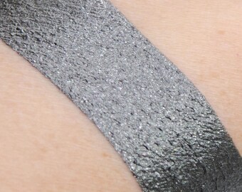 Op de afbeelding: Een close-up van een zilveren oogschaduw swatch op de huid. De oogschaduw heeft een metallic finish en een fijne shimmer.