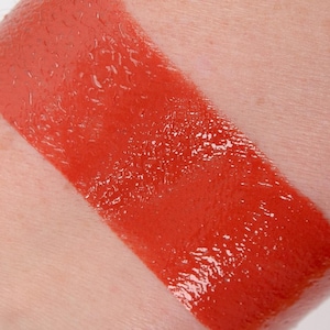 Puede incluir: Un primer plano de una muestra de lápiz labial de color rojo anaranjado brillante en el brazo de una persona. El lápiz labial tiene un acabado brillante.