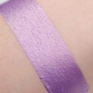 Pode incluir: Uma amostra de sombra para os olhos de cor roxa cintilante no braço de uma pessoa. A sombra para os olhos tem um acabamento metálico e é um tom de roxo claro.