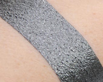 Op de afbeelding: Een close-up van een zilveren oogschaduw swatch op de huid. De oogschaduw heeft een metallic finish en een fijne shimmer.