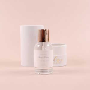 May include: A clear glass bottle with a gold cap and a white label that reads "Honey Grace Honey Blossom" is sitting on a white surface. The bottle is next to a white tube with the text "Honey Grace" in gold lettering.