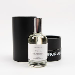 May include: A clear glass bottle of Connor Allen BOLD fragrance with a black and white label. The bottle is sitting on a black cylindrical box with the text "Connor Allen" printed on it.