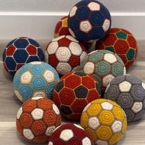 May include: A collection of spherical crochet balls featuring a geometric pattern of interlocking hexagons in various colors including navy blue, red, yellow, teal, gray, and cream. Each ball is constructed from yarn with a visible circular stitch.