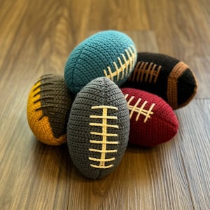 May include: Five crocheted footballs in different colors, including blue, brown, yellow, red, and gray. Each football has white stitching.