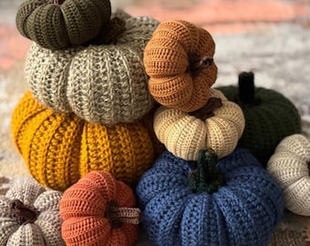 Puede incluir: Una colección de calabazas hechas a mano con ganchillo en varios colores, incluyendo naranja, amarillo, verde y azul. Las calabazas están apiladas, mostrando su diseño texturizado y acanalado y sus tallos marrones. Decoración de otoño.