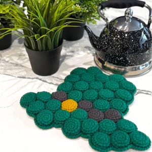 May include: A handmade, green crochet trivet with a flower design, featuring a yellow center and gray petals. A black and silver speckled tea kettle and small potted plants are in the background.