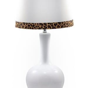 Può includere: Lampada da tavolo in ceramica bianca con paralume in tessuto bianco e bordatura con stampa leopardata marrone e nera. La lampada ha una base rotonda.