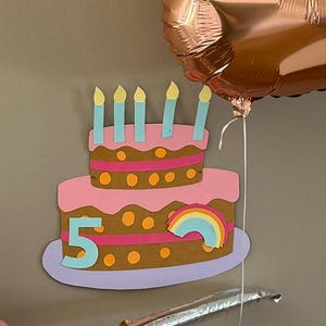 Könnte beinhalten: Ein zweistöckiger Kuchen in Rosa und Braun mit einem Regenbogen und der Zahl "5" darauf. Der Kuchen hat blaue Kerzen und ist mit orangefarbenen Punkten verziert.