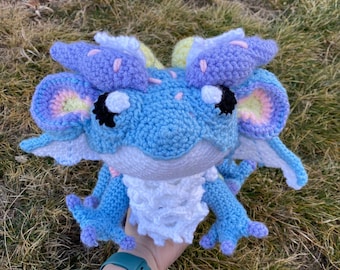 Puede incluir: Un dragón de ganchillo hecho a mano en tonos de azul, morado y blanco. El dragón tiene alas, orejas y pies grandes y detallados. Los ojos del dragón son negros y blancos. El dragón está sentado sobre un lecho de hierba.