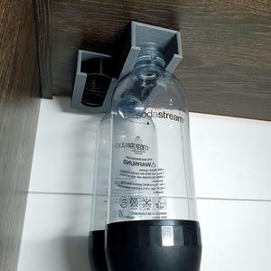 May include: Two clear plastic soda stream bottles with black caps are hanging from a gray plastic holder attached to a brown wooden surface. The bottles have a black label with the text "sodastream" and a warning label in a foreign language.