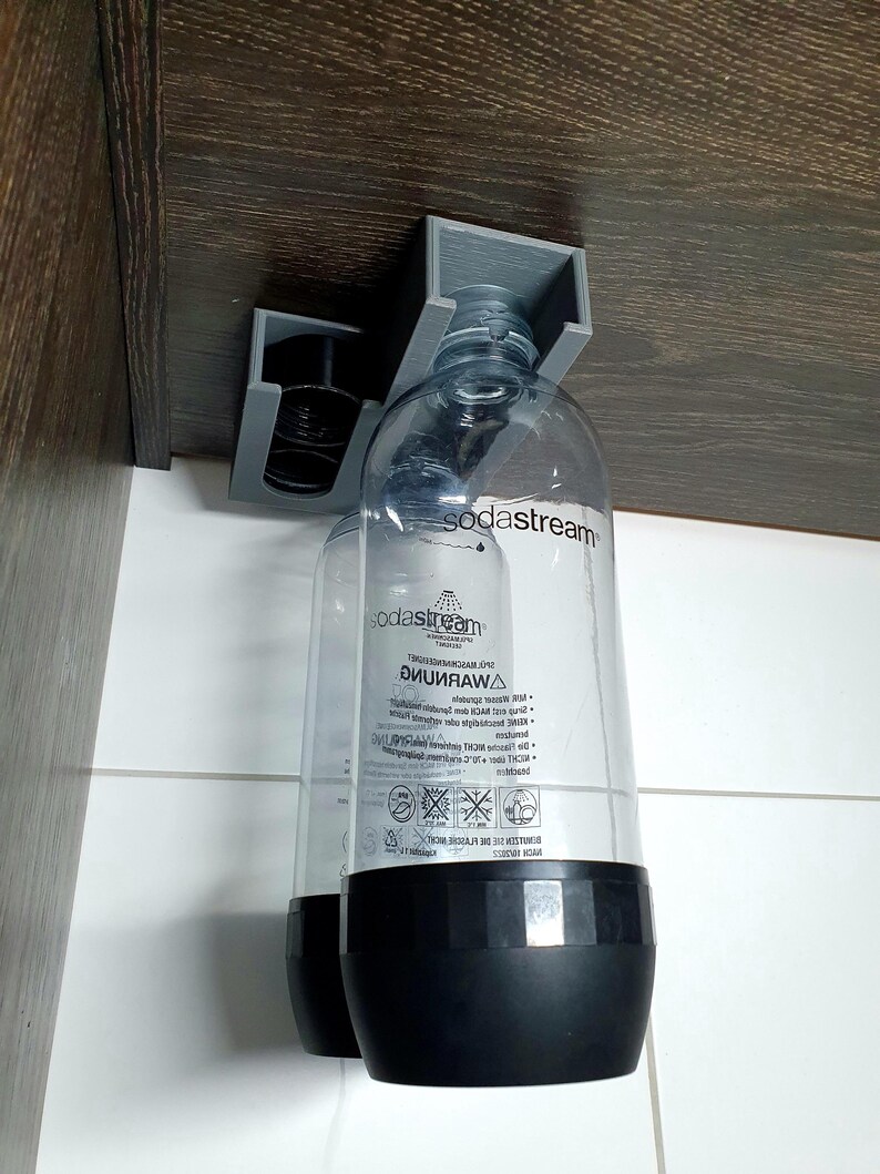 May include: Two clear plastic soda stream bottles with black caps are hanging from a gray plastic holder attached to a brown wooden surface. The bottles have a black label with the text "sodastream" and a warning label in a foreign language.