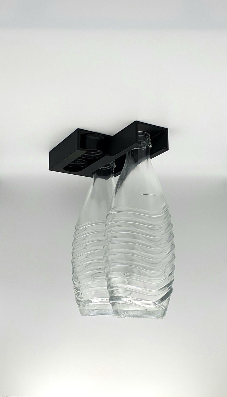 May include: Two clear glass bottles with textured surfaces are held upside down by a black plastic holder.