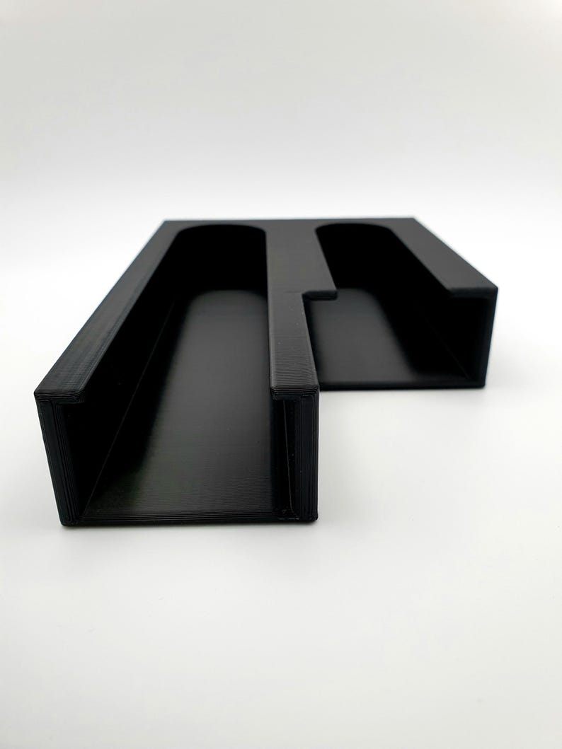 May include: Black plastic organizer with two slots for holding items. The organizer is designed to be mounted on a wall.