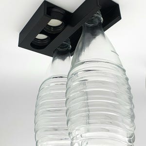 May include: A black metal bottle holder with two clear glass bottles hanging upside down. The bottles have a textured surface.