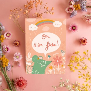 May include: A pink card with a green mouse wearing heart-shaped sunglasses and a speech bubble that says 'On s'en fiche!' The mouse is surrounded by flowers and a rainbow. The card is on a pink background with dried flowers.