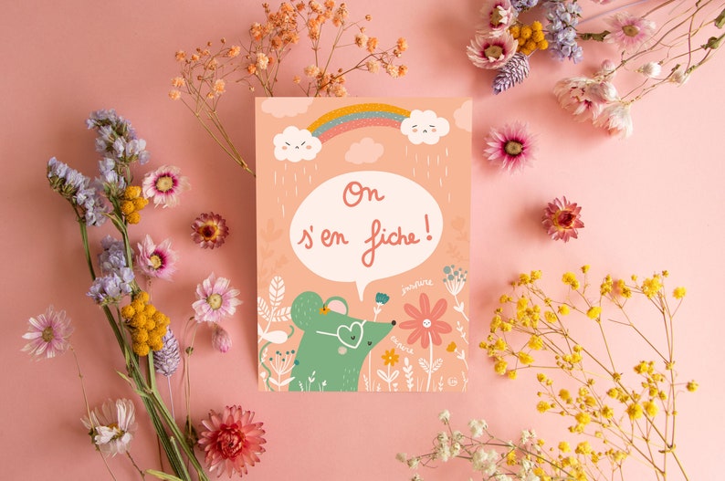 May include: A pink card with a green mouse wearing heart-shaped sunglasses and a speech bubble that says 'On s'en fiche!' The mouse is surrounded by flowers and a rainbow. The card is on a pink background with dried flowers.