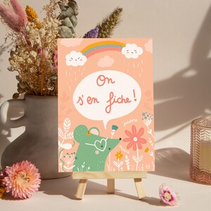 May include: A pink card with a green mouse wearing heart-shaped sunglasses and a speech bubble that says 'On s'en fiche!' The card is decorated with flowers, a rainbow, and clouds. The card is on a small wooden easel.