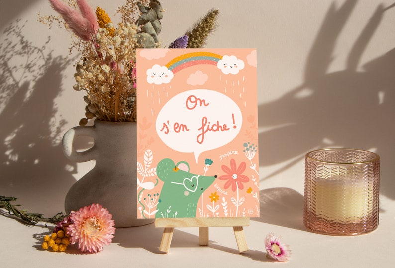 May include: A pink card with a green mouse wearing heart-shaped sunglasses and a speech bubble that says 'On s'en fiche!' The card is decorated with flowers, a rainbow, and clouds. The card is on a small wooden easel.