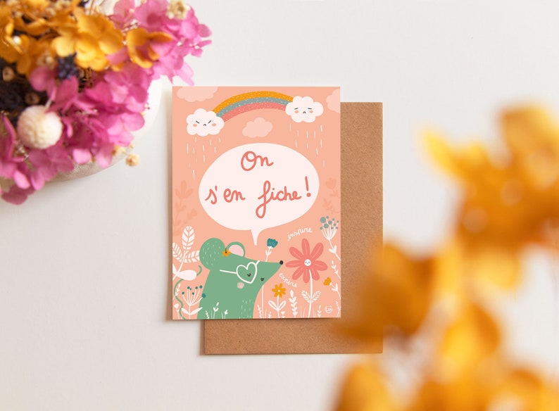 May include: A pink card with a green mouse wearing glasses and a heart-shaped nose. The mouse is smelling a pink flower with a smiley face. The card has a rainbow and clouds with rain drops. The text on the card reads 'On s'en fiche!'.