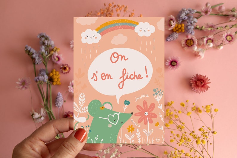May include: A pink card with a green mouse wearing sunglasses and a speech bubble that says 'On s'en fiche!' The card has a rainbow, clouds, and raindrops. The mouse is in a field of flowers with the words 'inspire' and 'explore' written on the flowers.