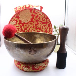 May include: A large, round, brass singing bowl with a red felt mallet and a wooden striker. The bowl is sitting on a red and gold patterned cushion. The bowl has a decorative symbol on the bottom.