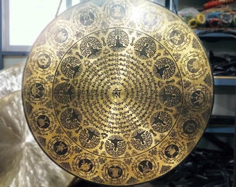 May include: A large, round, gold-colored gong with intricate black designs and lettering. The gong is hanging from a metal hook and has a raised, circular design in the center.