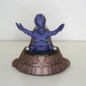 May include: Purple alien figure with a large head and outstretched arms sitting on a bronze throne with a circular base.