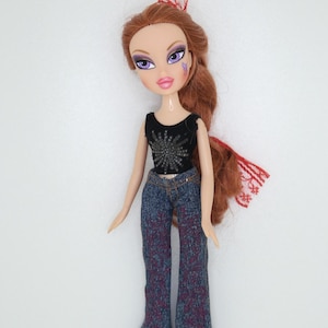 May include: A doll with long auburn hair styled with a red lace accessory. The doll wears a black sleeveless top with a silver starburst graphic, blue denim flared pants featuring a dark red floral pattern, and tan animal print pointed boots.