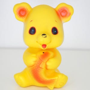 Pode incluir: Um brinquedo de urso de borracha amarelo vintage com detalhes laranja nas orelhas e uma língua vermelha. O urso está segurando um peixe laranja e vermelho. O brinquedo tem olhos pretos e está sentado.