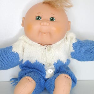 May include: A vintage baby doll with a bald head, green eyes, and a tuft of blonde hair. The doll is wearing a blue and white knitted outfit with a button-up sweater and matching trousers. The doll is sitting down.