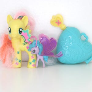 Può includere: Un pony giocattolo giallo con capelli rosa e arancioni e un pony viola più piccolo con ali di farfalla. È presente anche un contenitore blu a forma di cuore con una parte superiore a forma di chiave. I giocattoli sono decorati con motivi floreali e a farfalla.