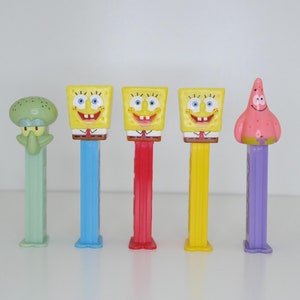 May include: Five colorful plastic character figurines on a white surface. The characters are from the cartoon Spongebob Squarepants. The characters are Spongebob, Patrick, Squidward, and two more Spongebobs. The figurines are attached to the tops of plastic dispensers.