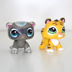 May include: Two small animal figurines. One is a grey raccoon-like figure with a mask and large eyes. The other is a yellow tiger with black stripes and green eyes. Both are plastic toys.