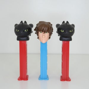 Puede incluir: Tres dispensadores de caramelos PEZ con personajes de la franquicia "Cómo entrenar a tu dragón". Dos dispensadores tienen cabezas de dragón negras sobre tallos rojos, y uno tiene una cabeza de personaje humano sobre un tallo azul.