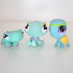 May include: Three toy turtle figurines in shades of blue and green. The turtles have large eyes and varying designs, including pink and yellow accents. One turtle has a yellow shell, and another has a green headband. The toys are on a white surface.
