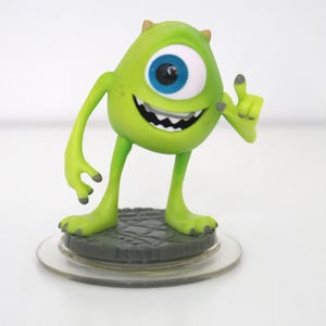 Op de afbeelding: Een limoengroen beeldje van het personage Mike Wazowski uit de film Monsters, Inc. De figuur heeft één groot blauw oog, een brede grijns en poseert met een opgestoken vinger. Het staat op een grijze basis met een doorzichtige plastic schijf.