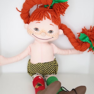以下が含まれることがあります： 緑のリボンで結ばれた赤い糸の三つ編みを持つ手作りのぼろきれ人形。人形は薄いピンク色の体、そばかすがあり、黄と黒のチェック柄のショーツ、赤と緑の靴下、茶色のブーツを履いています。人形は腕を広げて座っています。