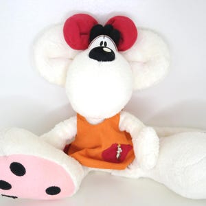 Könnte beinhalten: Ein Stofftier-Maus mit großen weißen Ohren und einer roten Schleife. Die Maus trägt ein orangefarbenes Kleid mit einer roten Tasche. Die Maus hat eine schwarze Nase und schwarze Augen. Die Füße der Maus sind rosa mit schwarzen Punkten.