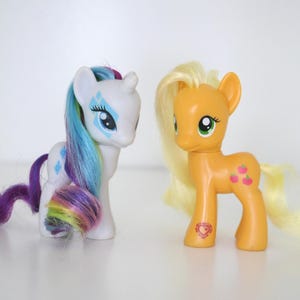 Può includere: Due pony giocattolo. Uno è bianco con criniera e coda arcobaleno e dettagli blu e viola. L'altro è arancione con criniera e coda gialle e segni di mela rosa. Entrambi hanno grandi occhi.