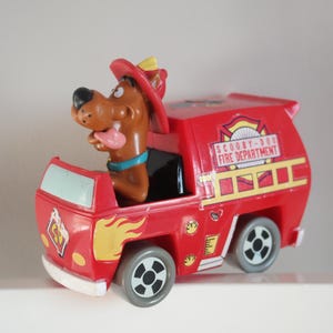 May include: A red Scooby-Doo Fire Department toy truck with a brown Scooby-Doo figure wearing a firefighter hat. The truck has yellow flames and the text "SCOOBY-DOO FIRE DEPARTMENT". The toy has gray wheels.