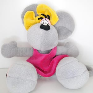 Könnte beinhalten: Ein graues Plüschmaus-Spielzeug mit einer gelben Schleife, einem rosa Kleid und schwarzen Augen. Das Stofftier hat große graue Ohren und sitzt. Das Spielzeug ist für Kinder konzipiert.