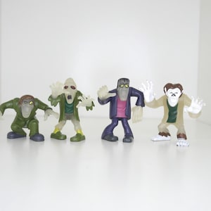 May include: Four small monster figurines in various colors. The figures include a green zombie, a Frankenstein's monster, and a werewolf. The figures are standing on a white surface.